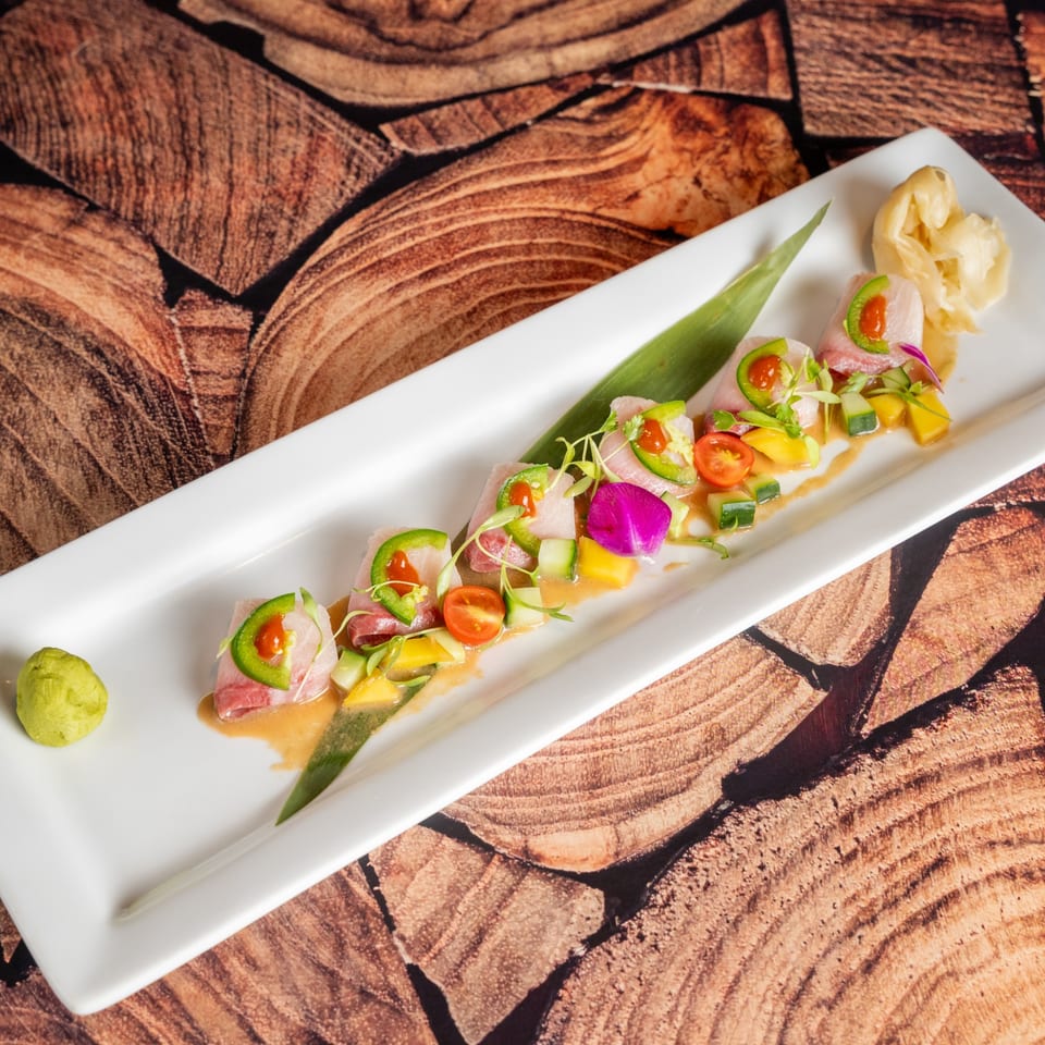 A long white plate holds a row of tuna sashimi with jalapeños, cherry tomatoes, and mango.